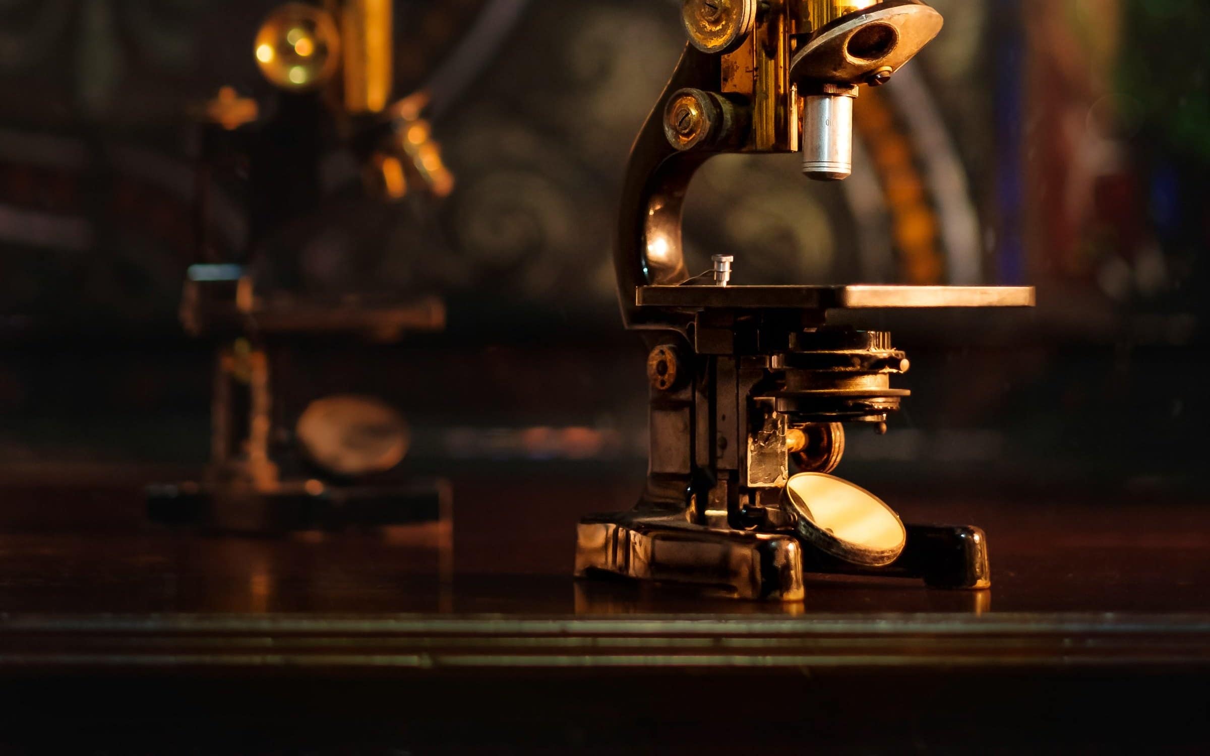 Two antique brass microscopes on a dark surface, lit warmly from one side.