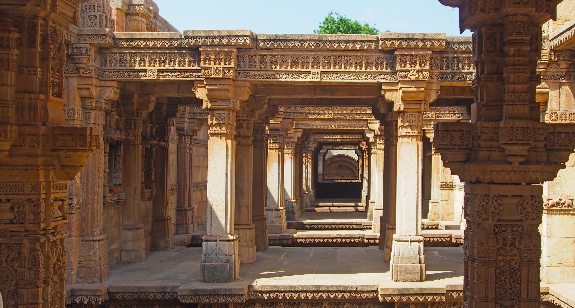 Carved stone pillars and beams of a stepwell, opening onto tiers of steps descending into shadow.