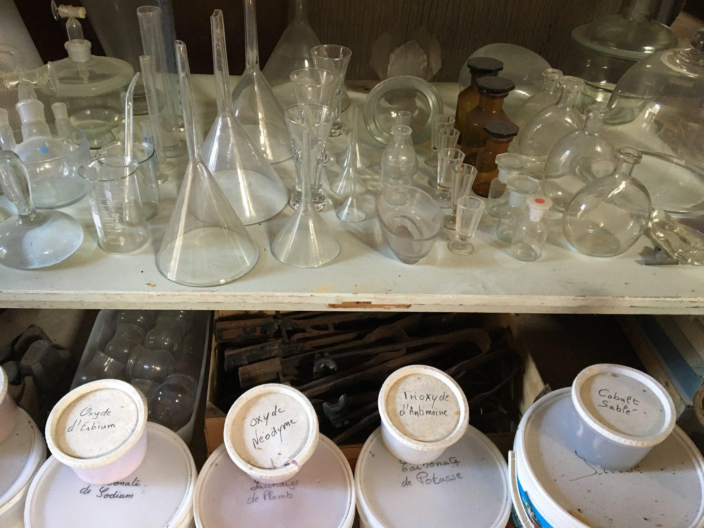 Shelves of laboratory glassware — funnels, flasks and measuring vessels — above a row of labelled ceramic reagent jars.