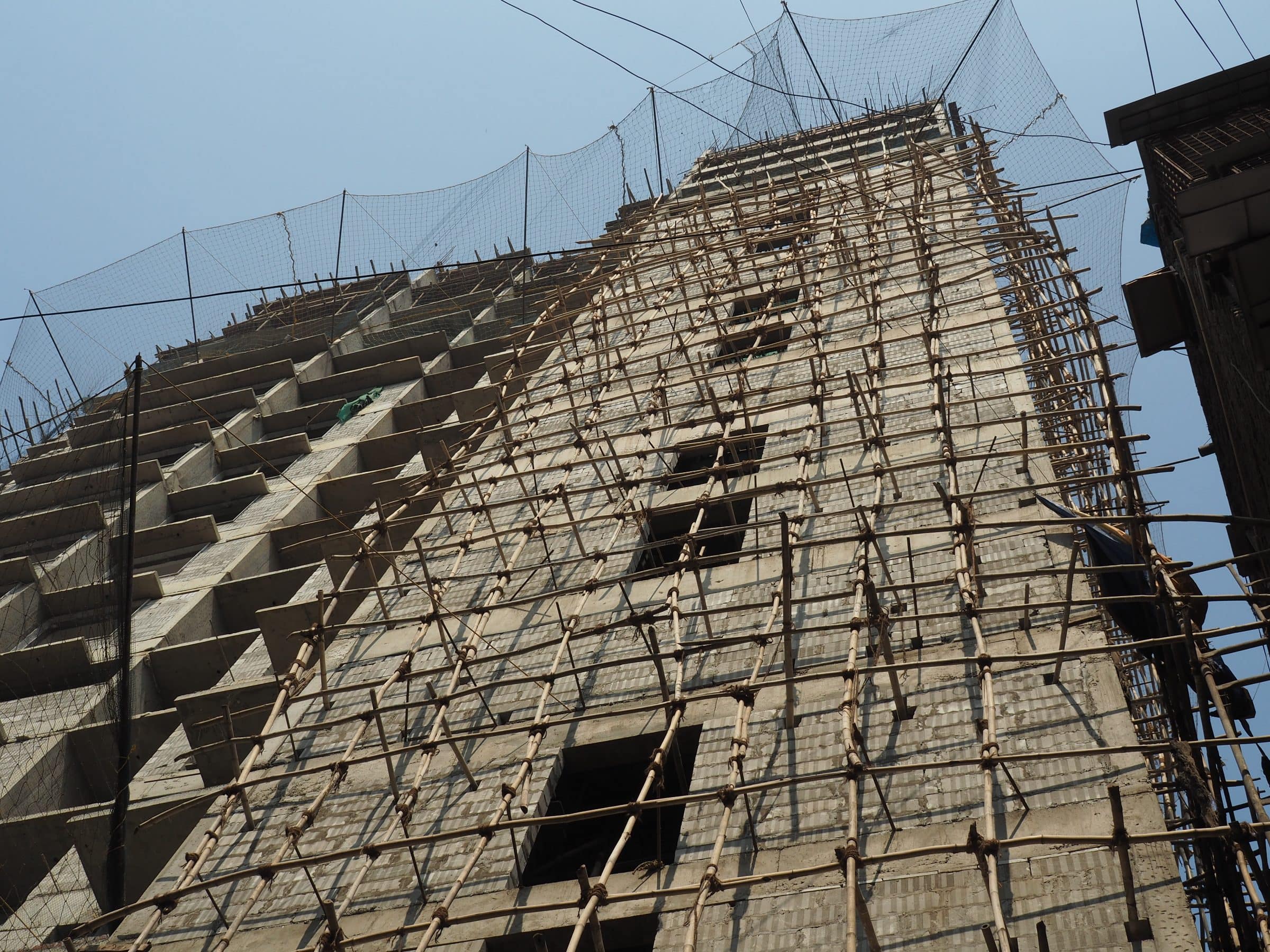 Bamboo scaffolding lashed in a lattice across the face of a tall concrete building under construction, seen from below against a pale sky.